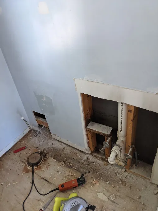 The wall has been cut away, revealing pipes for a sink and toilet
